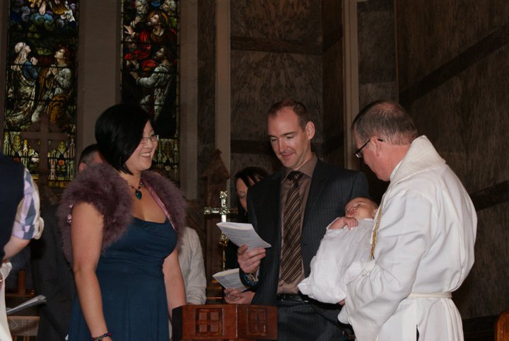 A celebratory baptism ceremony in a church, featuring a smiling woman in a blue dress and a fur shawl holding her child, while a man in a suit stands beside her. A priest is holding the baby, and stained glass windows are visible in the background.